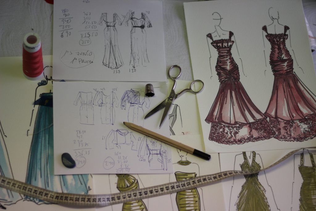 Varios dibujos de vestidos están sobre una mesa con tijeras y un lápiz.
