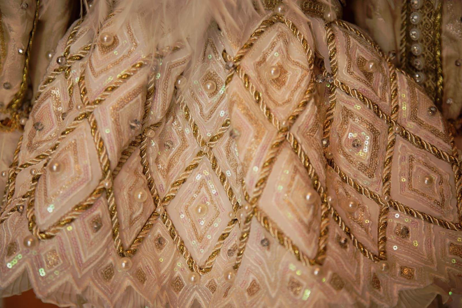 Primer plano de una tela adornada de color crema con patrones de diamantes delineados en cuerda dorada y adornada con cuentas y lentejuelas.