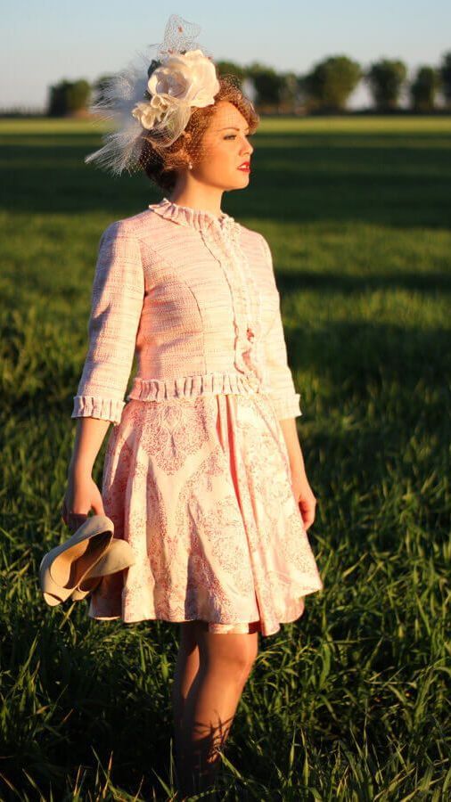 Una mujer con un vestido rosa está parada en un campo sosteniendo un sombrero.