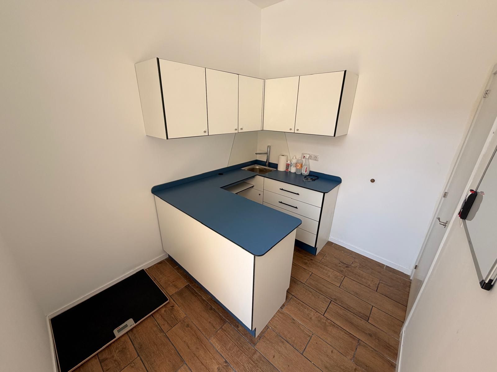 Pequeña cocina con armarios blancos, encimeras azules y suelo de imitación madera en una luminosa habitación de esquina.