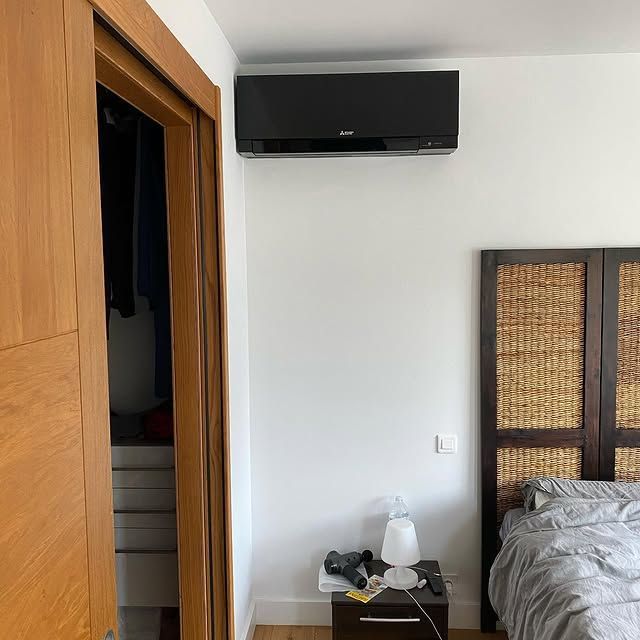 Chambre avec murs blancs, télévision noire fixée au mur et lit à côté d'une porte de placard en bois.
