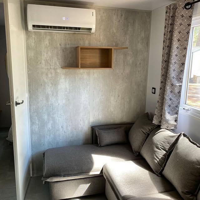 Petit salon gris avec canapé d'angle, étagère murale et climatiseur près de la fenêtre.