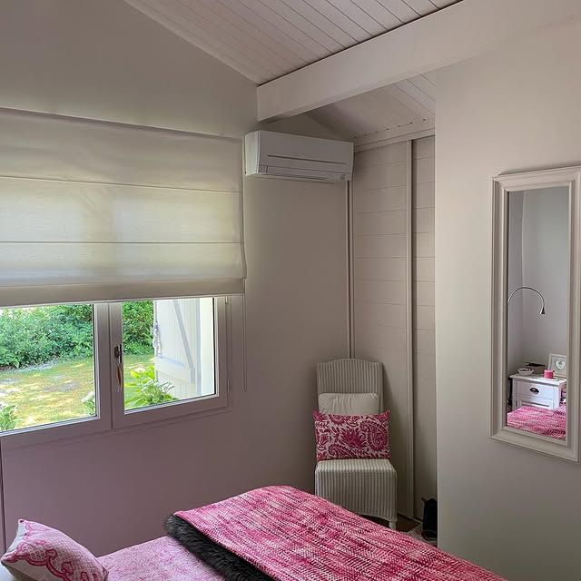 Chambre confortable avec literie rose, stores, miroir mural et petite chaise sous un climatiseur mural.