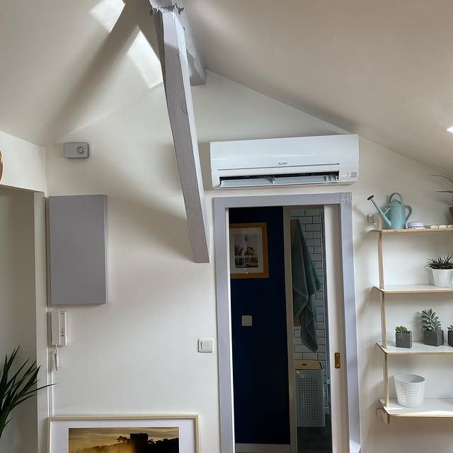 Chambre mansardée confortable avec plafond en pente, climatiseur mural, porte ouverte et étagères blanches avec plantes.