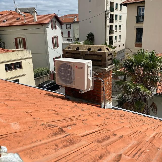 Vue depuis le toit avec des tuiles orange, un climatiseur mural et des immeubles d'appartements à proximité sous un ciel nuageux.