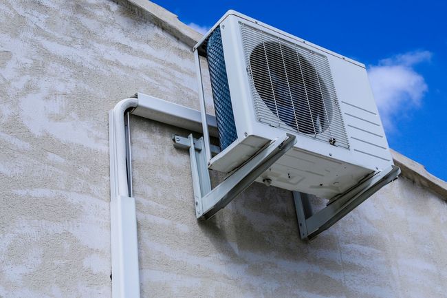 Unité de climatisation extérieure fixée sur un mur en stuc, se détachant sur un ciel bleu.