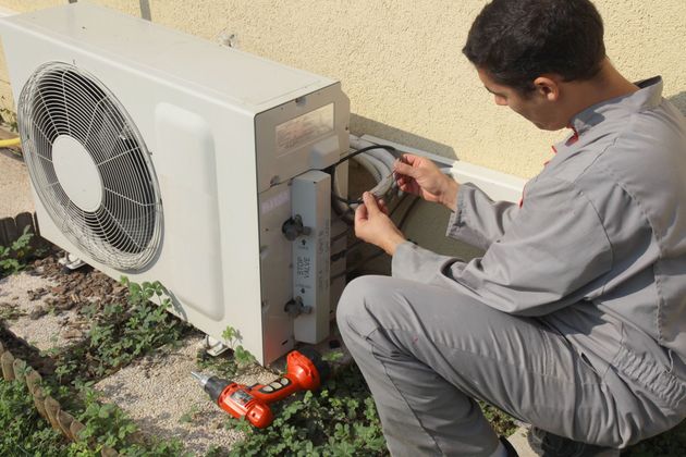 Un technicien répare un climatiseur extérieur adossé à un mur, ses outils posés au sol.