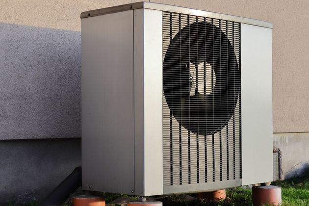 Unité de climatisation extérieure contre un mur en stuc, avec une grille de ventilateur noire et des panneaux beiges.