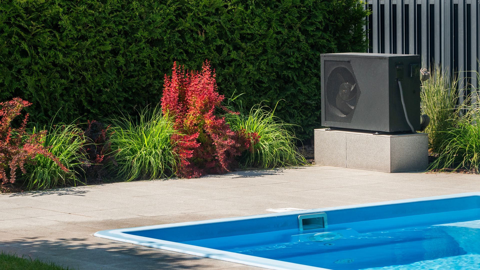 Piscine dans le jardin avec eau bleue, terrasse en béton et grande unité de climatisation extérieure, le tout entouré d'un aménagement paysager verdoyant.