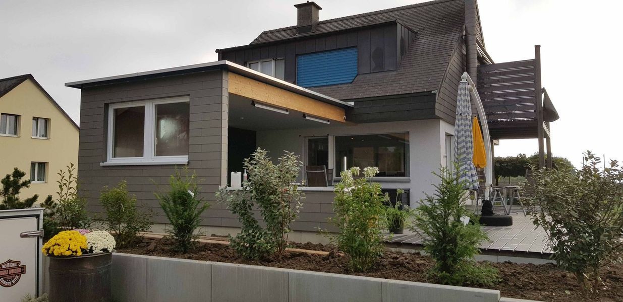 Ein großes Haus mit einer Veranda und vielen Fenstern ist von Bäumen und Büschen umgeben.