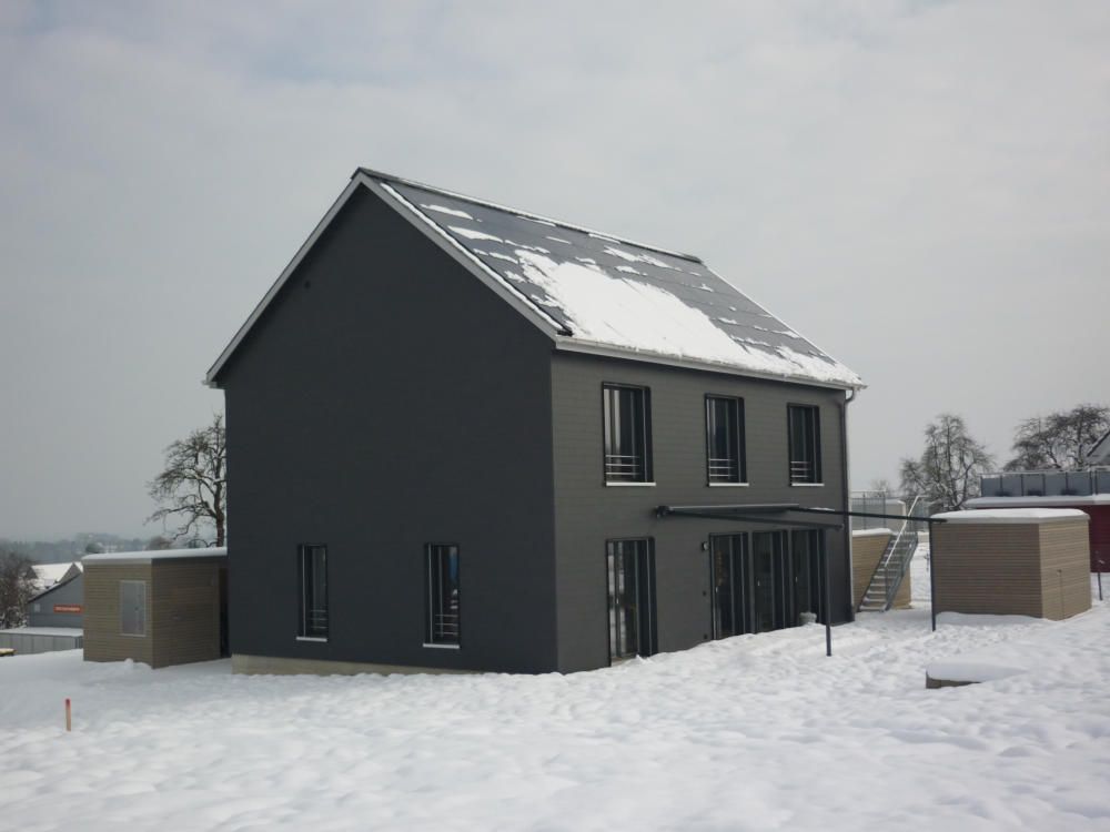Ein schwarzes Haus mit Schnee auf dem Dach ist von Schnee umgeben. Foto von der Mannhart Holzbau AG
