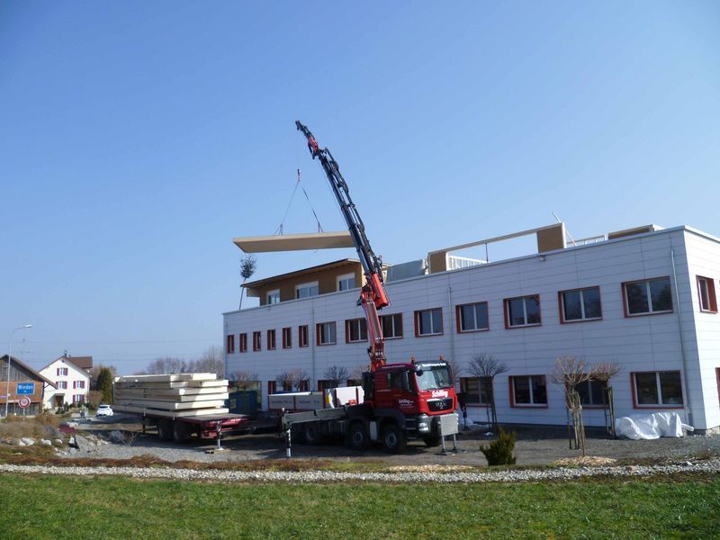 Ein roter Lkw mit einem daran befestigten Kran steht vor einem Gebäude. Foto von der Mannhart Holzbau AG