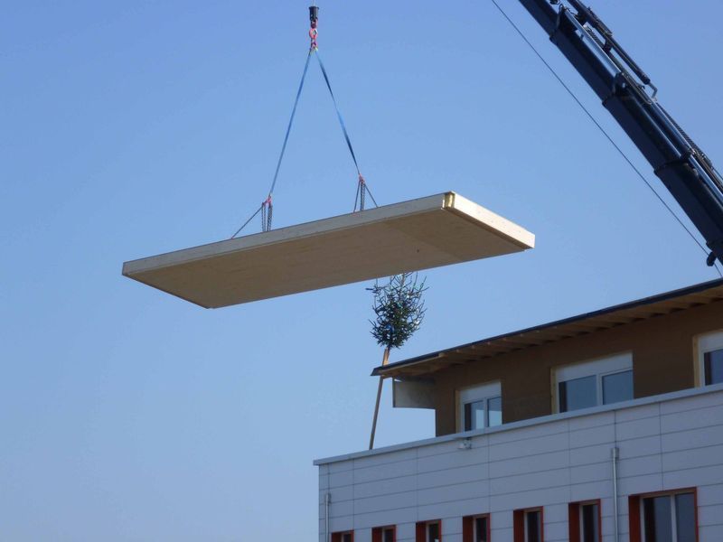 Ein Kran hebt ein Stück Holz über ein Gebäude. Foto von der Mannhart Holzbau AG