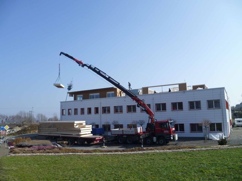 Ein roter Lkw mit einem daran befestigten Kran steht vor einem Gebäude. Foto von der Mannhart Holzbau AG