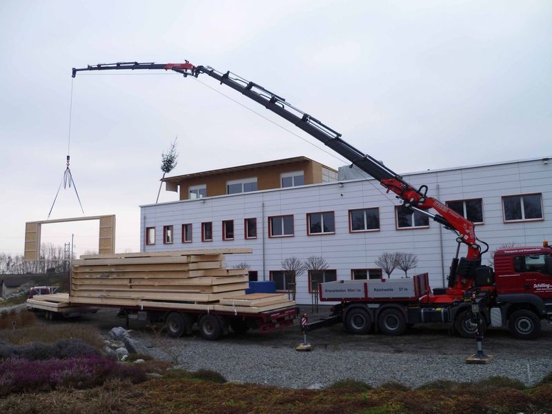 Ein Kran hebt ein Stück Holz vor einem Gebäude an. Foto von der Mannhart Holzbau AG
