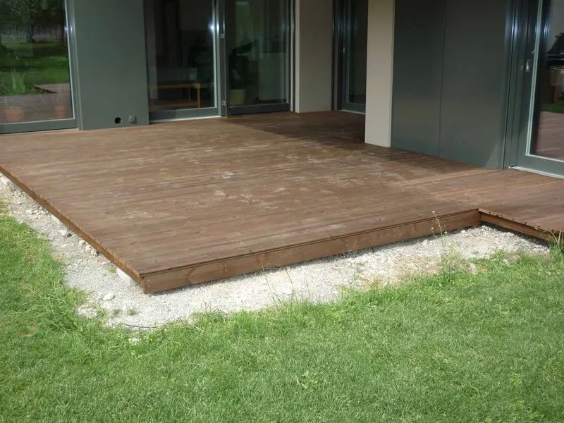 Vor einem Haus liegt auf einem üppigen grünen Rasen eine Holzterrasse. Foto von der Mannhart Holzbau AG