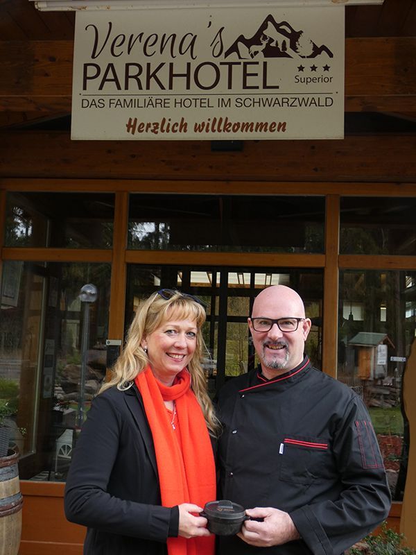 Ein Paar steht unter einem Hotelschild, die Frau mit rotem Schal, der Mann in Kochuniform. Schild: „Verena’s Parkhotel“.