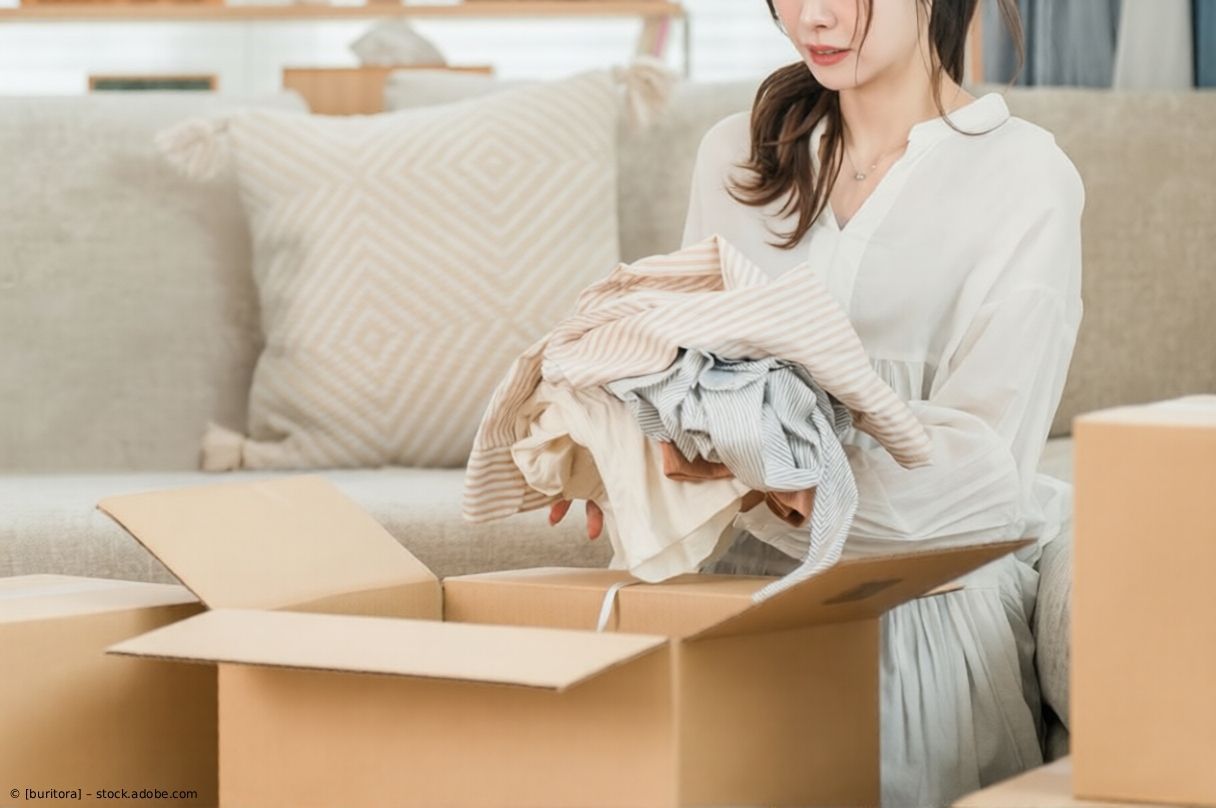 Frau packt Kleidung auf einem Sofa in einem hellen Wohnzimmer in einen Karton.
