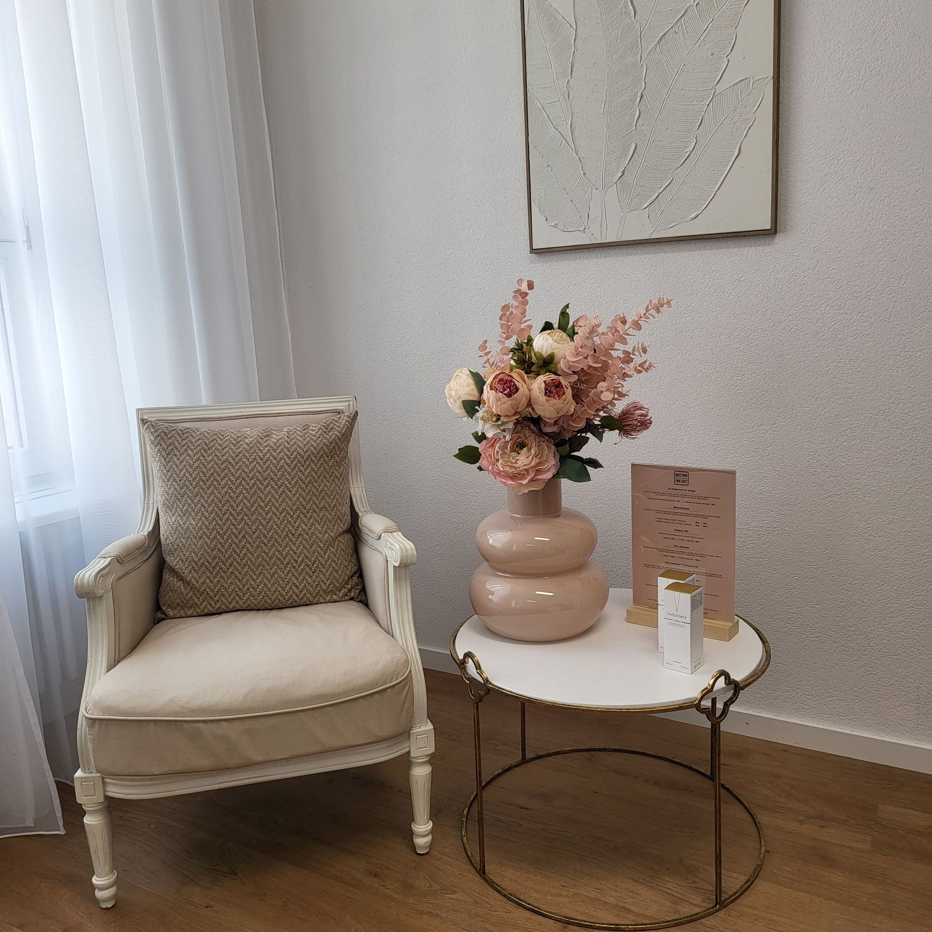 Un fauteuil beige confortable est placé à côté d'une table ronde blanche ornée de fleurs roses et de bougies dans une pièce douce et lumineuse.