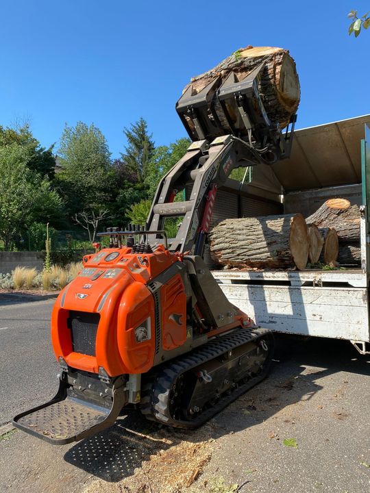 Un robot de démolition orange télécommandé décharge des gravats d'un camion sur un chantier extérieur ensoleillé.