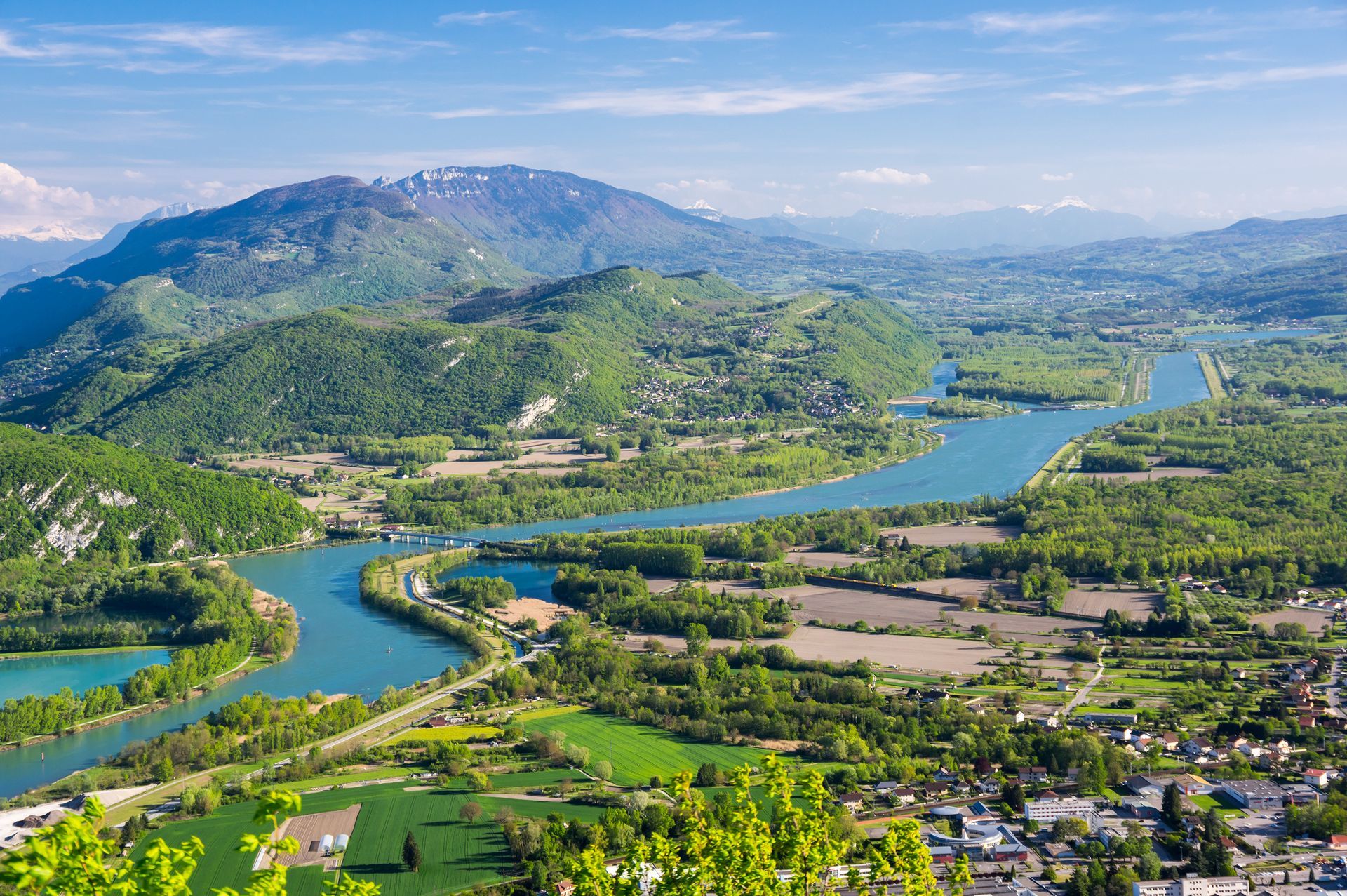 Une large rivière serpente à travers une vallée verdoyante, entourée de montagnes, de terres agricoles et d'une petite ville, sous un ciel d'un bleu limpide.