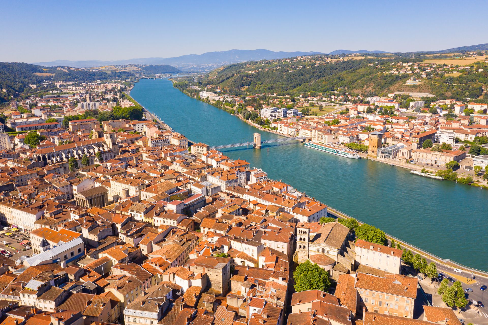 Vue aérienne d'une ville riveraine avec des bâtiments aux toits orange, un large fleuve bleu et des collines à l'horizon.