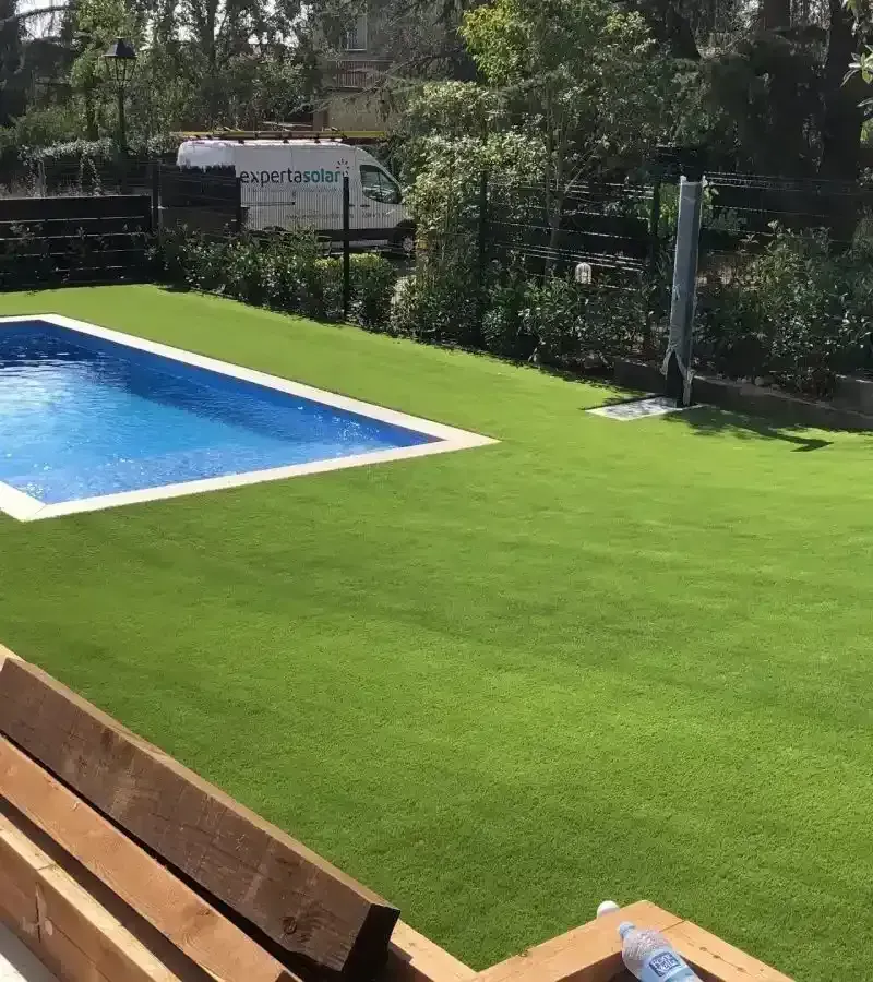 Piscina en el patio trasero con césped artificial de color verde brillante y un banco de madera en primer plano.