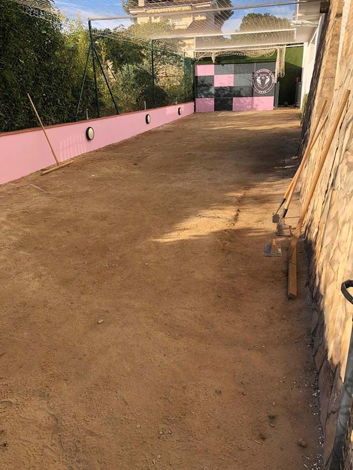 Un camino de tierra vacío junto a un muro y una puerta de color rosa, con un edificio y árboles al fondo.