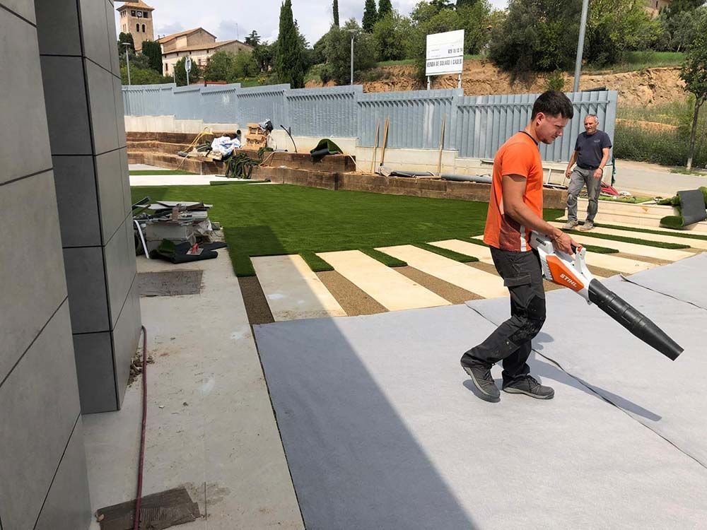 Un trabajador utiliza un soplador de hojas en un patio de concreto junto a césped recién cortado y losas para caminar al aire libre.