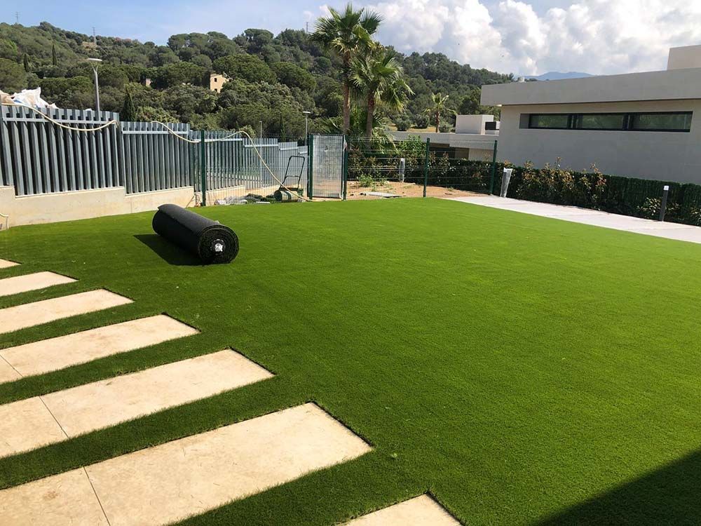 Patio trasero moderno con césped artificial, losas para caminar y un objeto cilíndrico negro cerca de una ladera vallada.