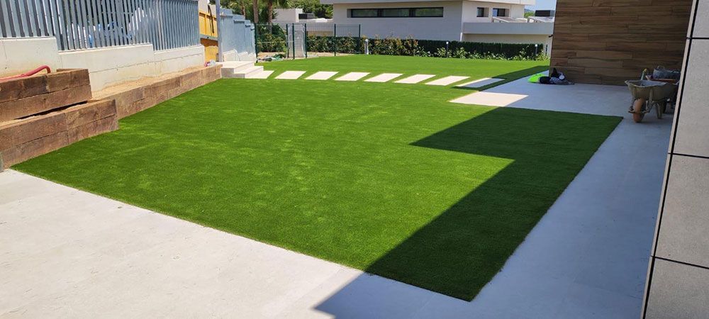 Pequeño patio trasero con césped artificial de color verde brillante, bordeado de hormigón y una terraza de madera.