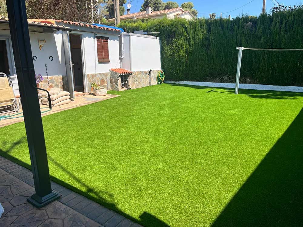 Patio trasero con césped artificial de color verde brillante, un pequeño patio y un seto junto a la casa.