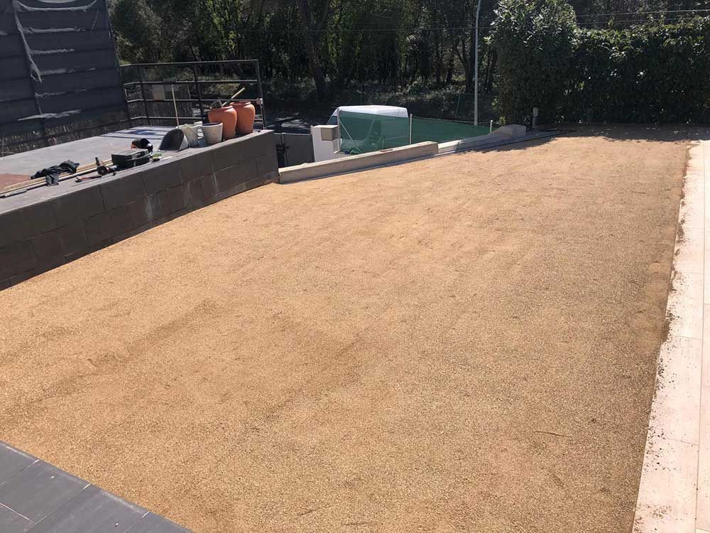 Patio exterior con una superficie de grava o arena de color beige recién extendida, junto a un muro de contención bajo y una valla.