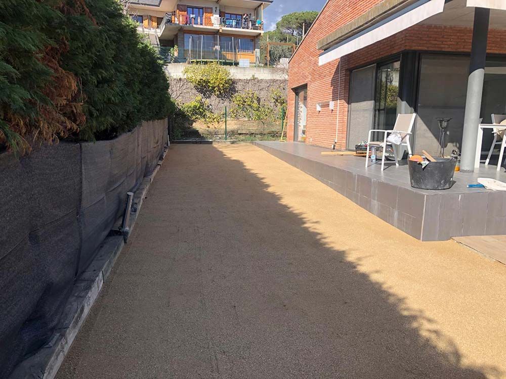 Patio trasero soleado con camino de grava, casa de ladrillo y sillas junto a una terraza cubierta.