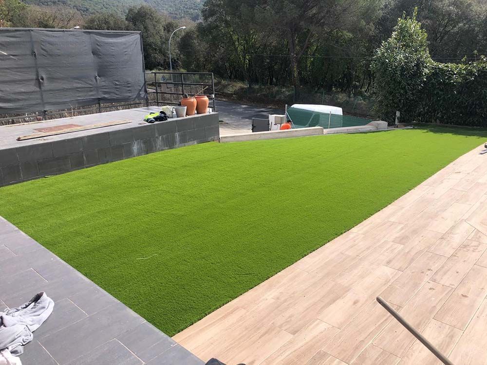 Patio trasero con césped artificial de color verde brillante, terraza de madera y patio de piedra en construcción.