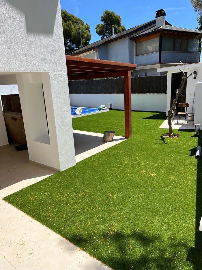 Patio trasero con césped verde, casa blanca, pérgola y una piscina inflable azul bajo el sol.