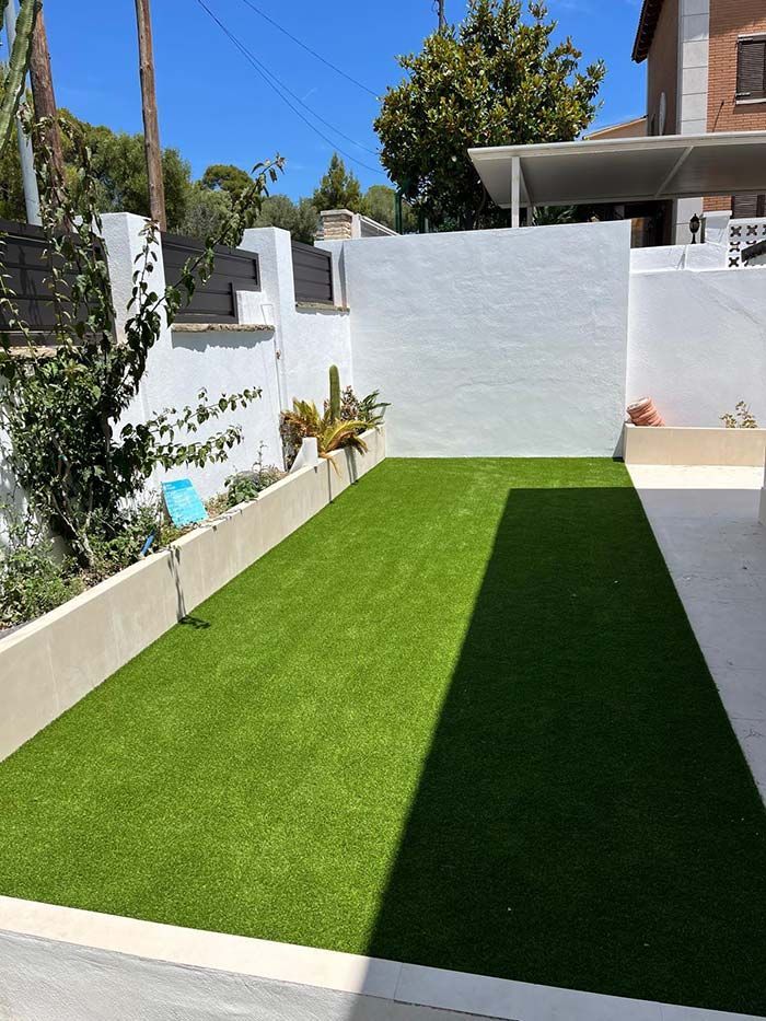 Pequeño patio trasero con césped verde brillante, paredes blancas y patio bañado por el sol; una larga sombra se extiende sobre el césped.