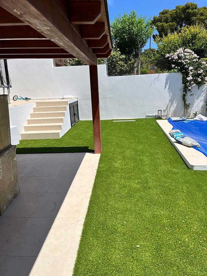 Patio trasero con césped verde, pared blanca, escaleras y área de estar techada.