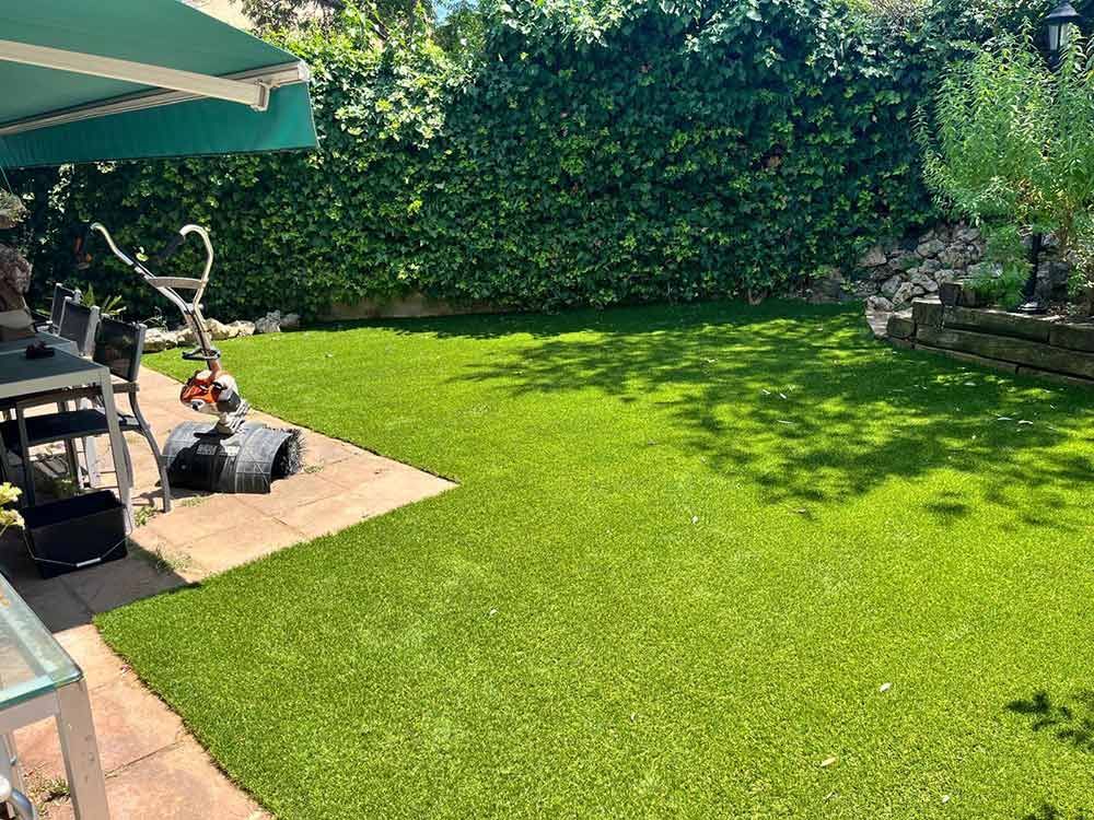 Patio trasero soleado con césped verde exuberante, sombrilla de patio y una pequeña zona de estar junto a una valla bordeada de setos.