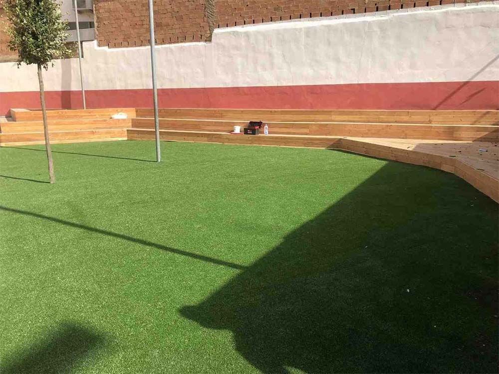 Patio con césped verde, muro de ladrillo, sombra y un pequeño perro blanco cerca del muro del fondo.