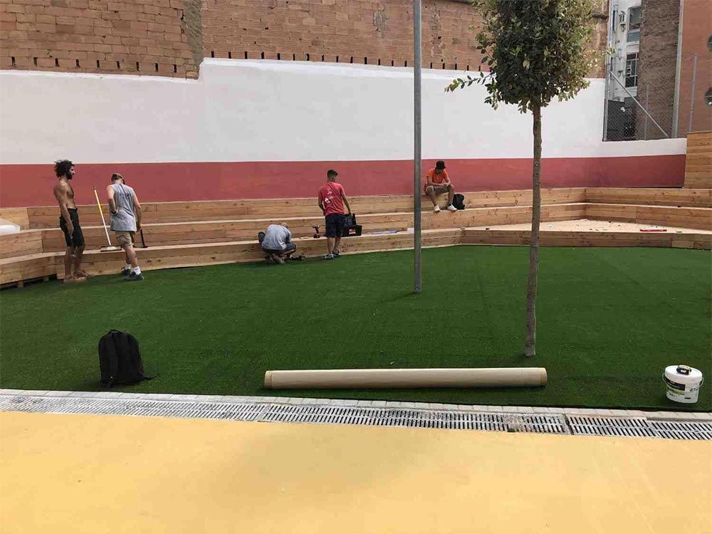 Personas trabajando en un pequeño patio ajardinado con césped verde, bancos de madera y un muro de ladrillos como telón de fondo.