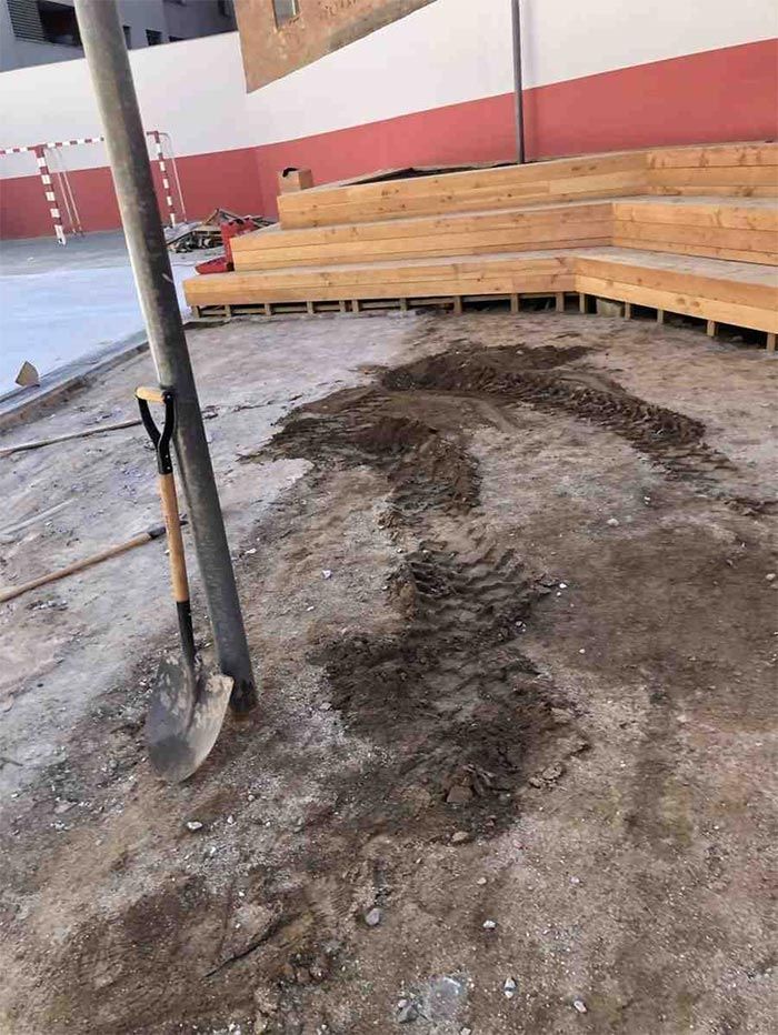 Suelo de tierra dañado con una zanja abierta junto a bancos de madera apilados en una habitación parecida a un gimnasio.