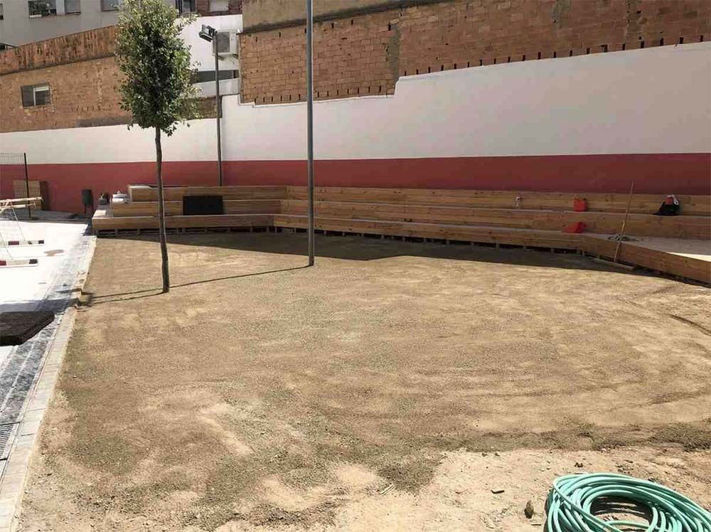 Patio exterior con banco curvo de ladrillo, un pequeño árbol y una manguera verde enrollada sobre terreno arenoso.