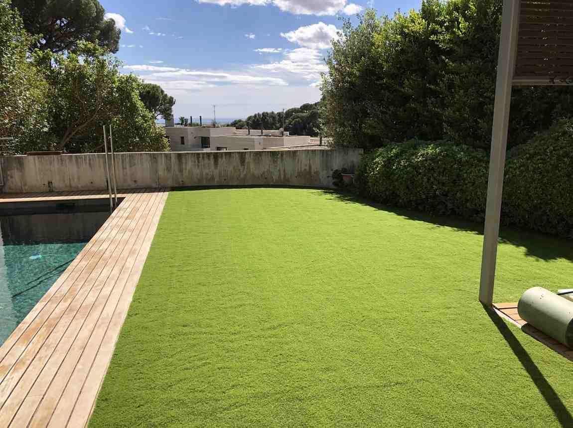 Patio trasero con césped artificial, borde de piscina y terraza de madera bajo un cielo soleado.