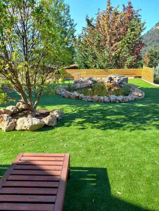 Patio trasero soleado con césped verde, árboles, jardinera de piedra y banco de madera en primer plano.