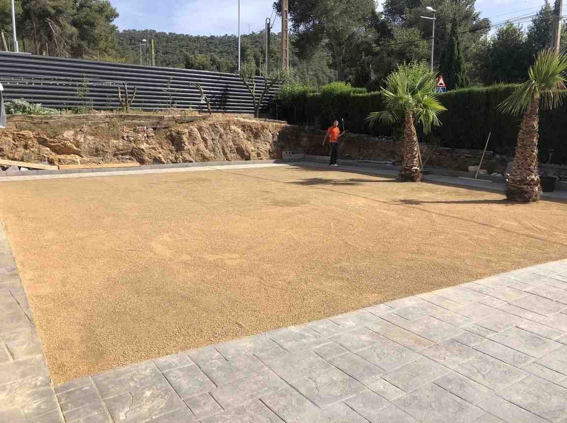 Patio soleado pavimentado con césped seco, palmeras y una persona vestida de naranja al fondo.