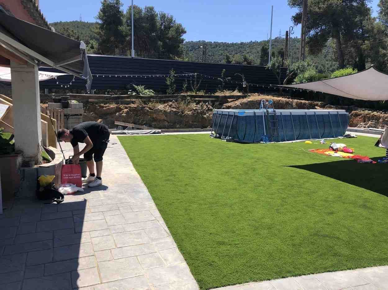 Una persona se inclina sobre objetos en un patio junto a un césped artificial de color verde brillante y una zona de piscina cubierta de color azul.