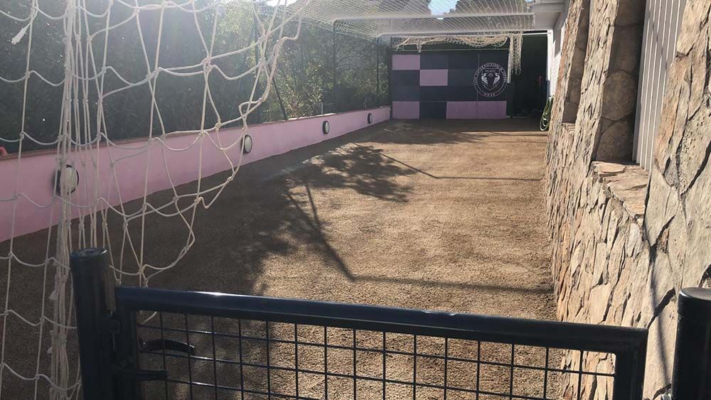 Patio de tierra bañado por el sol con paredes rosas y grises, una red en el techo y una puerta negra en primer plano.