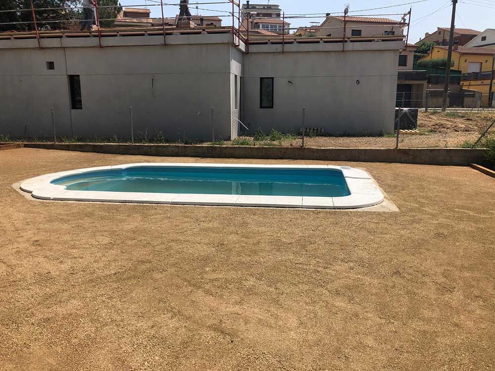 Pequeña piscina rectangular en el patio trasero con agua azul sobre césped seco junto a un edificio gris.