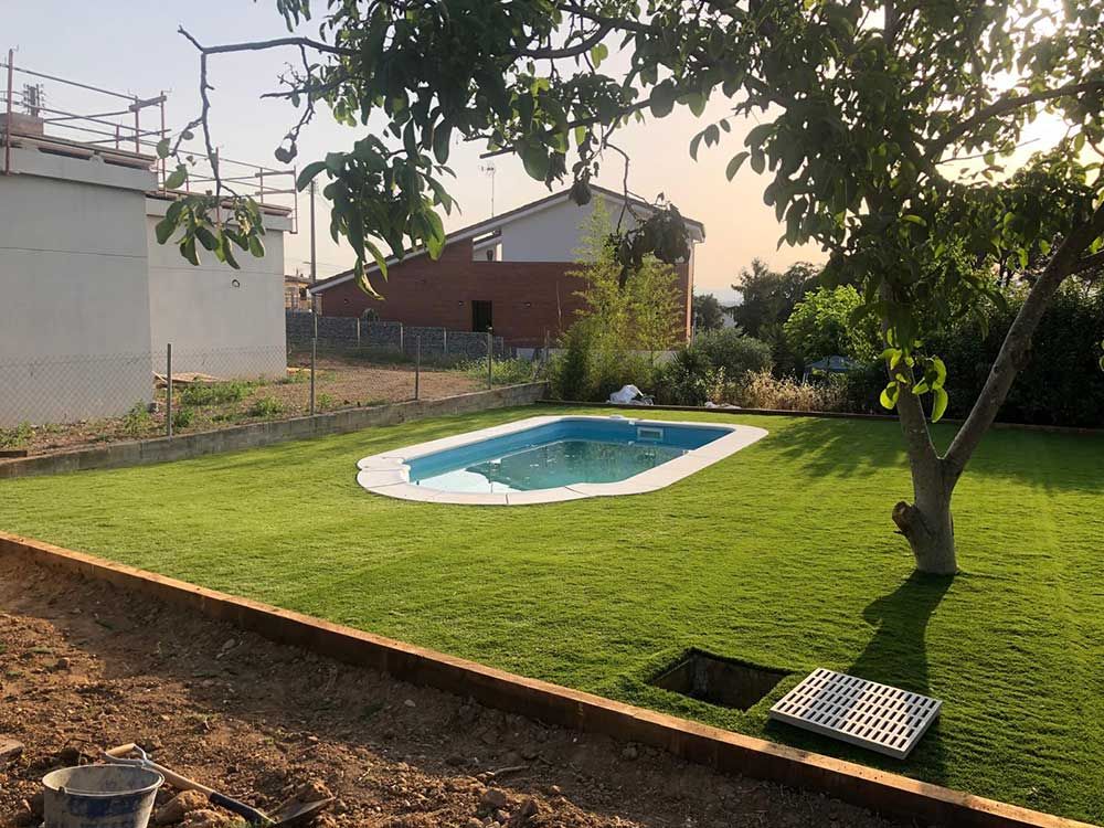 Patio trasero con una pequeña piscina azul, césped verde, árboles y una casa al fondo.